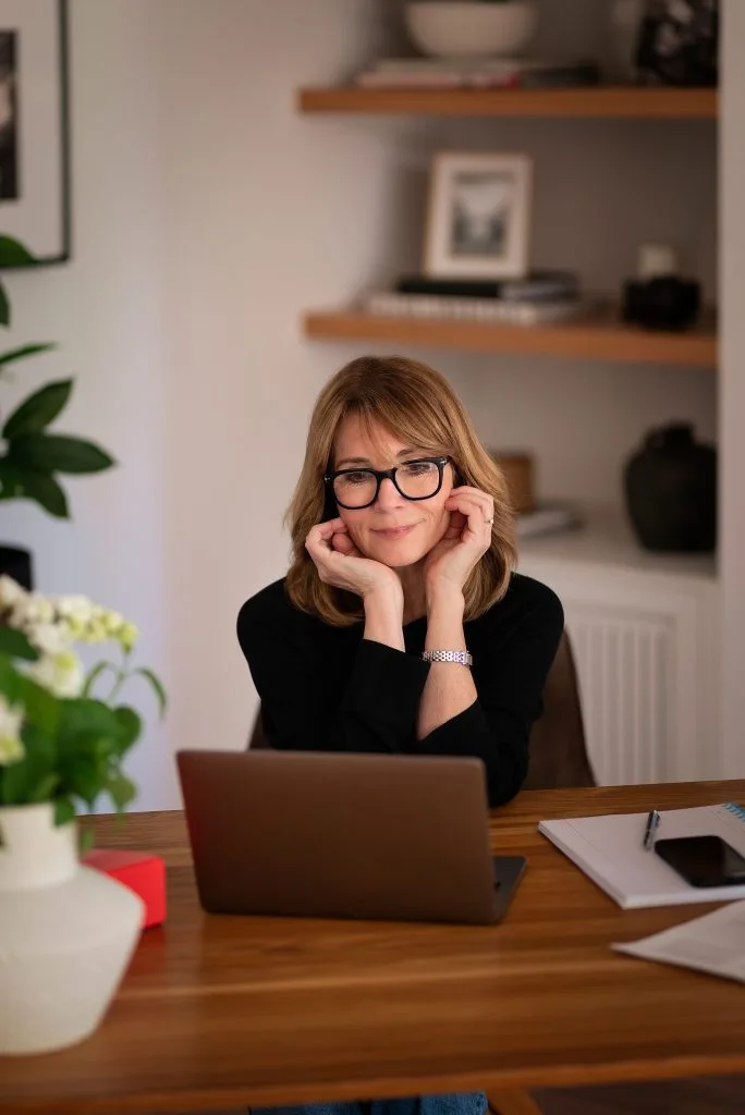 Woman with glasses smiling at laptop while working at home office desk