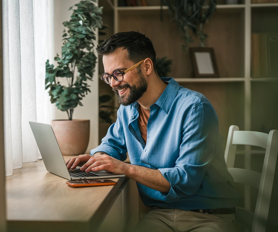 Man in blue shirt smiling while typing on laptop at desk with potted plant and shelving