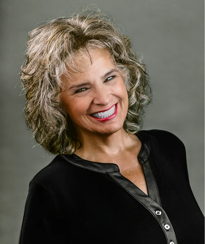 Woman with curly gray hair smiling, wearing black blazer with satin trim