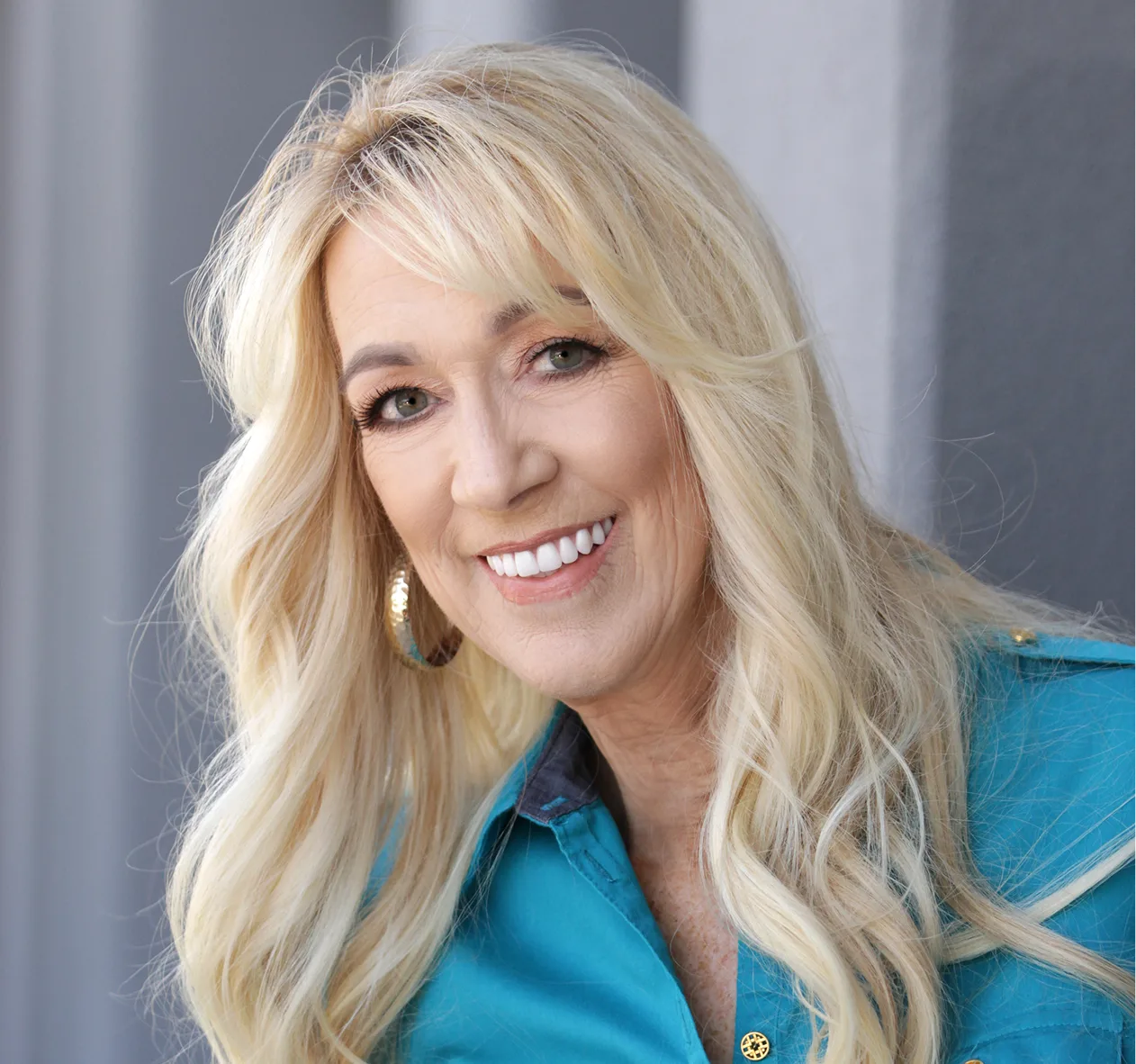 Woman with blonde wavy hair smiling, wearing turquoise shirt and gold hoop earrings