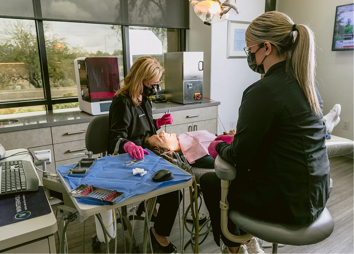 Dentist treating patient in dental chair while assistant observes in modern operatory with large windows