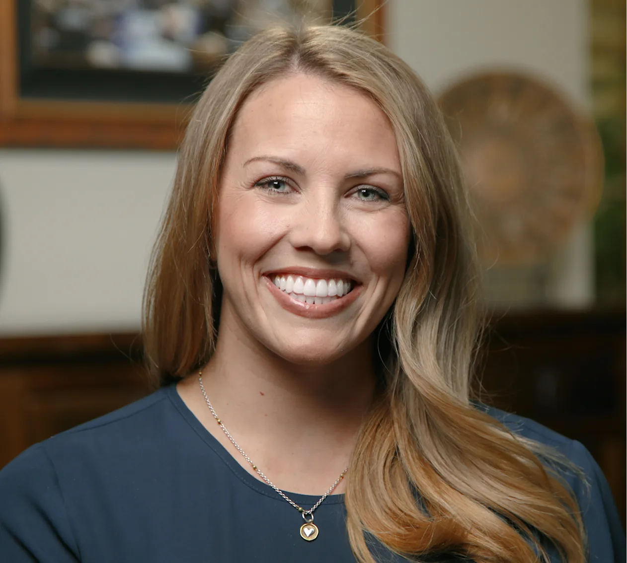 Woman with long brown hair smiling at camera wearing dark blue top and pendant necklace