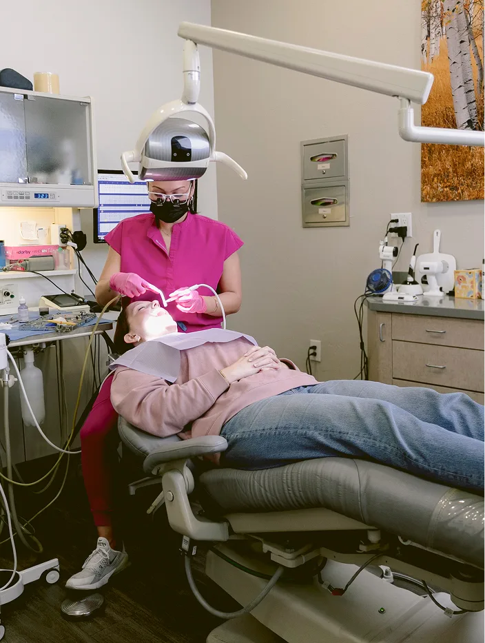 Dentist in magenta scrubs examining patient's teeth in treatment chair with overhead light and dental instruments