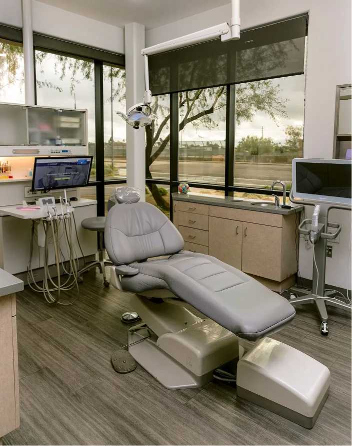 Modern dental operatory with patient chair, overhead light, computer workstations, and large windows overlooking trees