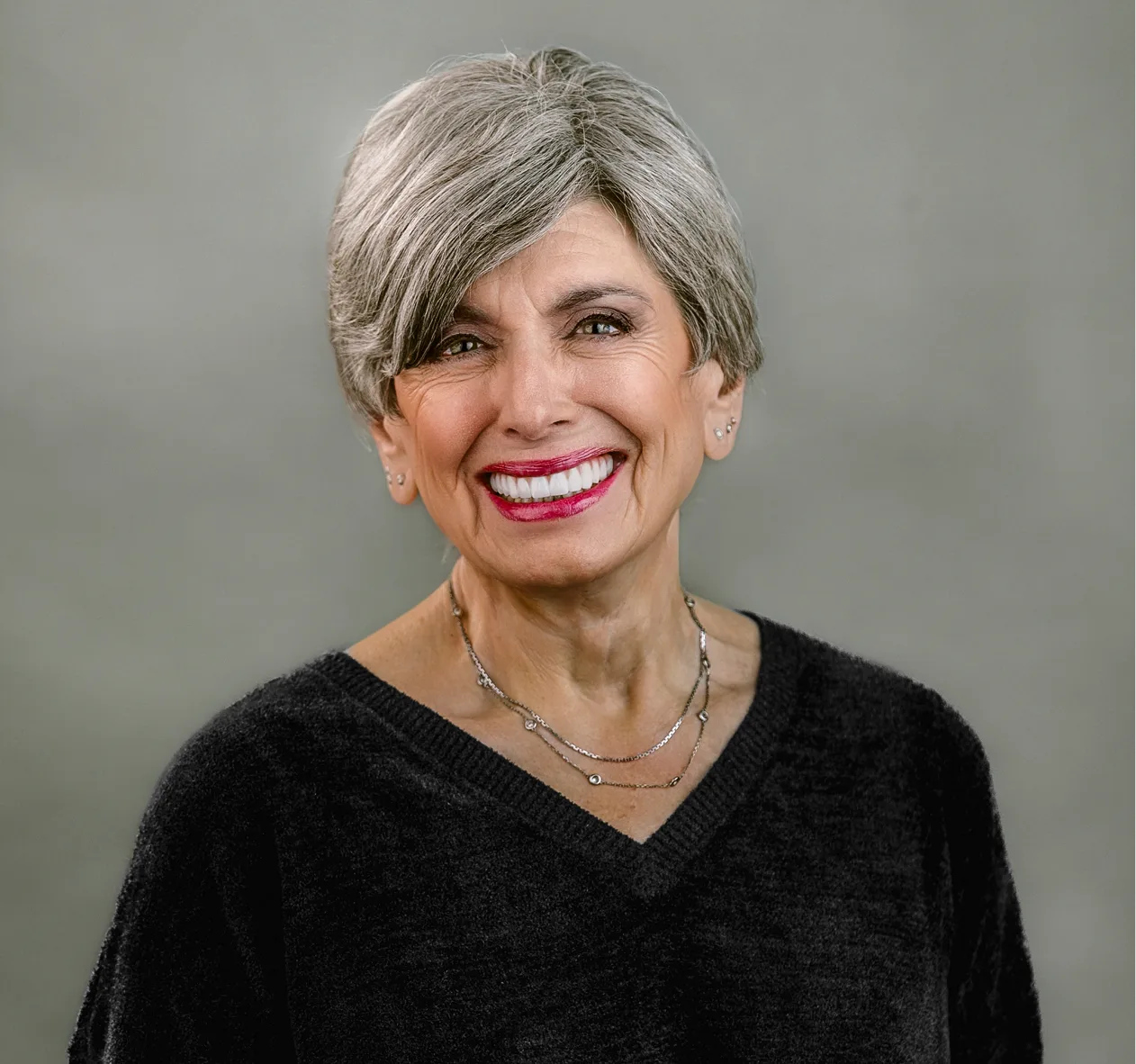 Woman with gray hair and red lipstick smiling at camera against gray background