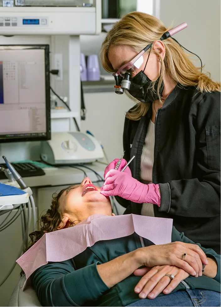 Dentist wearing magnification loupes and pink gloves examining patient's teeth in operatory