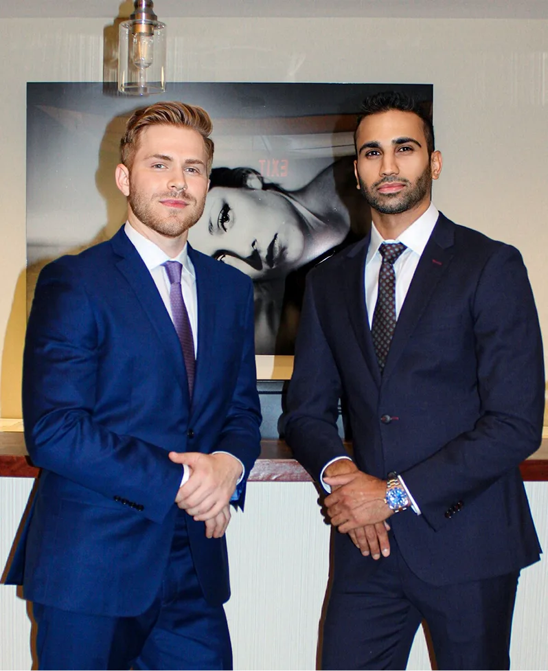 Two men in dark business suits shake hands in front of a black and white portrait on an office wall