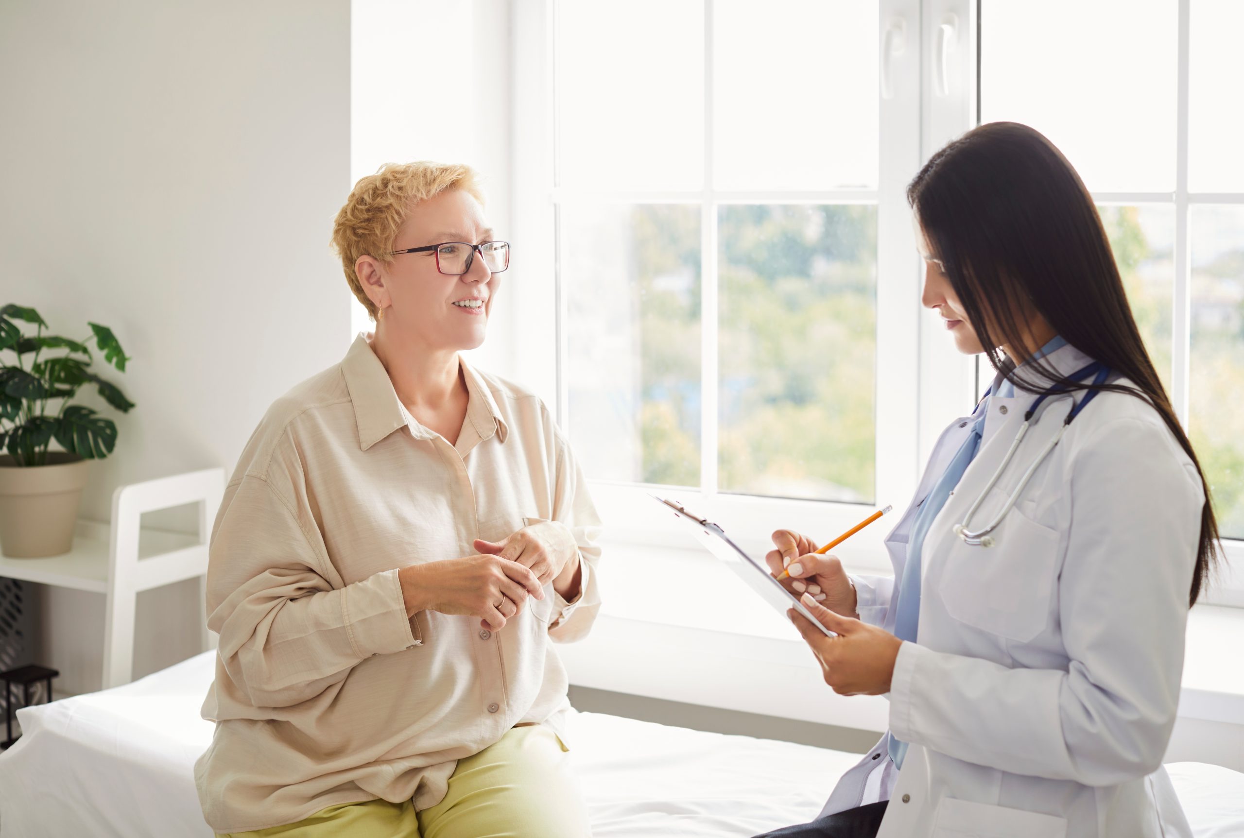 Healthcare provider in blue coat with stethoscope consulting with patient seated by a bright window