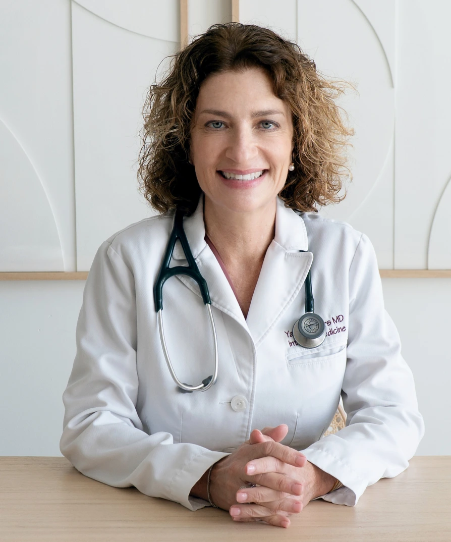 Female physician in white lab coat with stethoscope seated at desk, hands clasped, smiling
